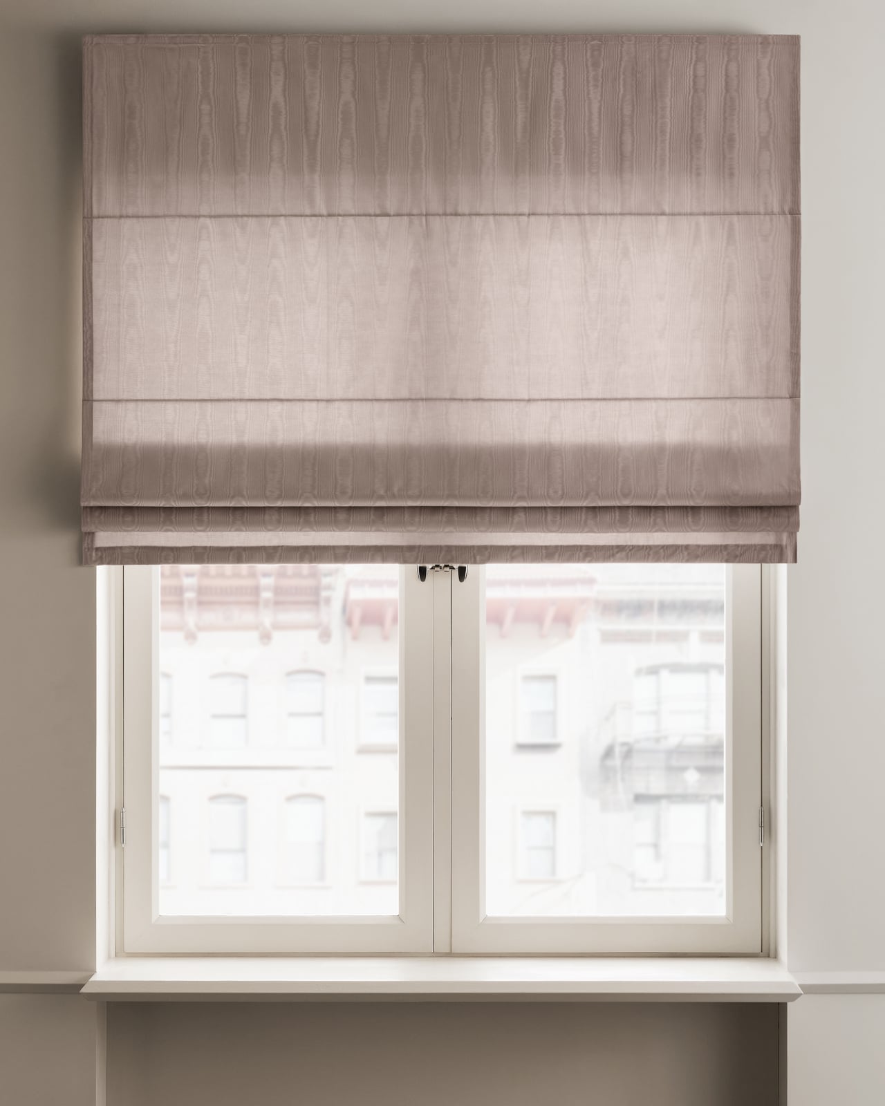 The Moire Roman Shade - Blackout Lining - Lavender in Lavender mounted over a white window framing an urban view.