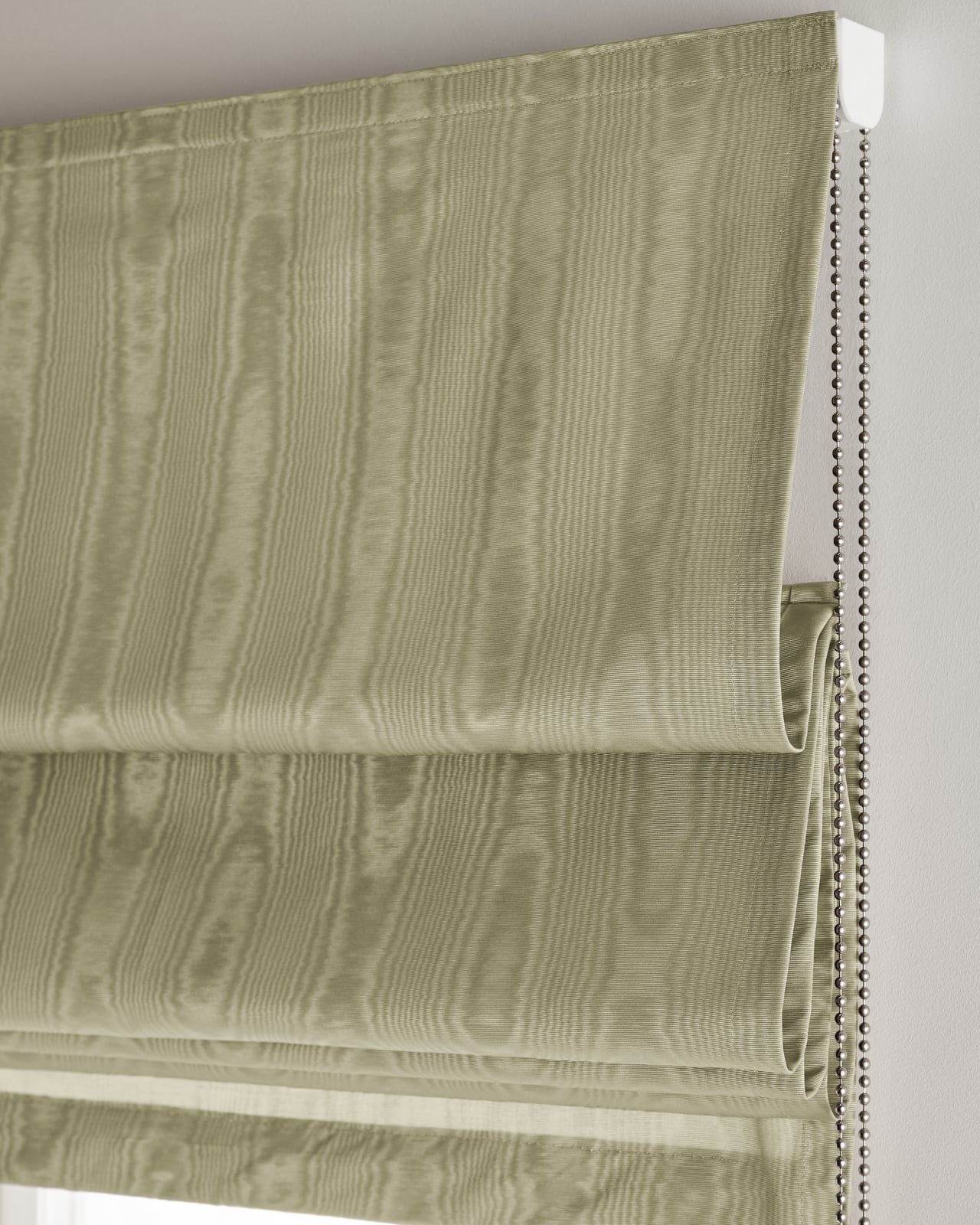 The Moire Roman Shade - Blackout Lining - Pistachio shown over a window, highlighting its silvery green moire pattern.