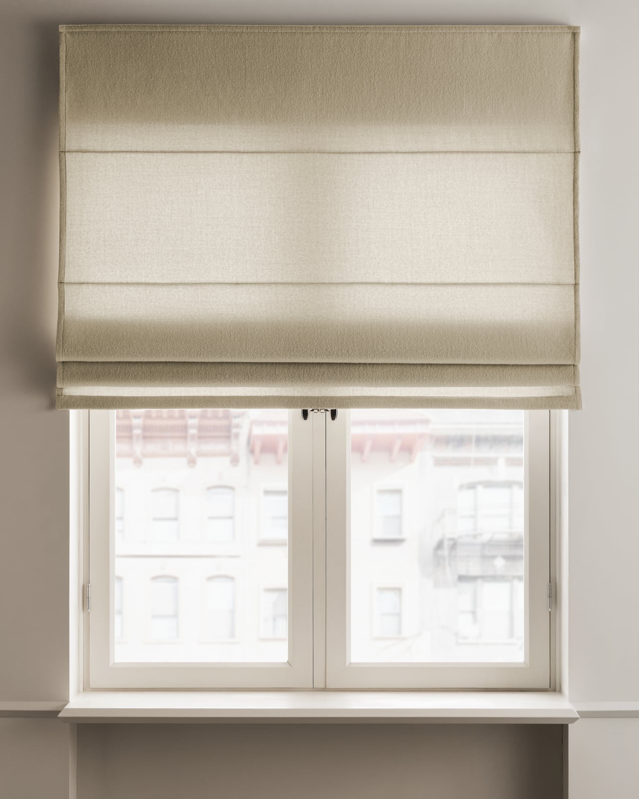 The Shelter Roman Shade in Sand, shown mounted over a window in a warm beige boiled wool fabric with horizontal folds.